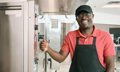 Black male foodservice operator wearing glasses, a red polo t-shirt, black apron and black hat poses in school kitchen and smiles while holding oven handle.