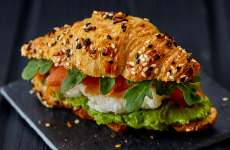 Croissant sandwich dusted with sesame seeds filled with smoked salmon, avocado, egg whites, and arugula on a black plate.