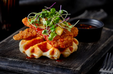 Biscuit waffle topped with breaded chicken, spicy honey, and arugula on black wooden board platter.
