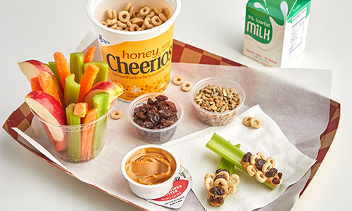Lunch tray with apple wedges, carrot and celery sticks, Honey Cheerios cereal cup, milk carton, raisins, sunflower seeds, peanut butter and “Bees on a Branch” recipe.