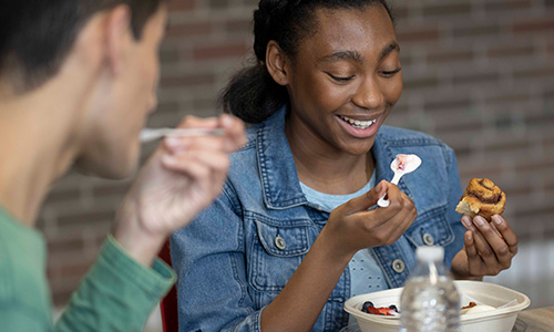 Female student smiling and raising a spoonful of yogurt with berries to her mouth, cinnamon roll in the other hand, with a male student eating in foreground.