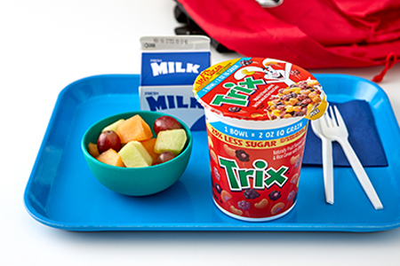 A cereal cup of Trix, an assorted serving of fruit, and a carton of milk sit on a blue lunch tray with a plastic fork and spoon.