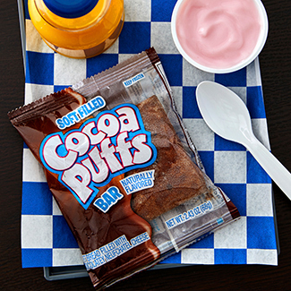Soft Filled Cocoa Puffs Bar placed on blue and white checkered place mat with a serving of yogurt and orange juice. Plastic spoon.