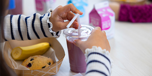 Hands of a female K-12 student inserting a straw into a yogurt smoothie cup with a muffin top and banana in a paperboard container with milk carton in the background on a table with other students.