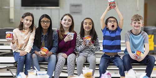 Group of six K-12 students sitting on a cafeteria table bench holding cup Lucky Charms cereal cups smiling.