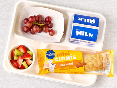 Cafeteria tray with container of red grapes, a carton of milk, a cup of sliced strawberries and kiwi next to a package of Pillsbury Mini Cinnis Caramel flavor.