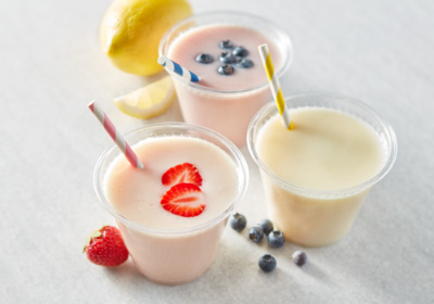 Three smoothies in plastic cups with straws. One garnished with sliced strawberries, another garnished with blueberries and one lemon smoothie without garnish.