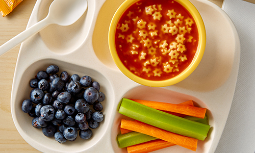 Beige lunch tray with plastic spoon, blueberries, soup with small noodles, celery and carrot sticks.