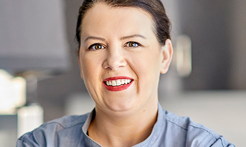 Headshot of Chef Heather Swan. Middle aged, light complexion with red lipstick and brown hair pulled back. Wearing grey chef’s coat. 