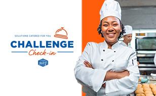 Challenge check-in text in blue and orange on a white background. Icon of a concierge bell in orange. Black female chef in white chef’s coat and hat smiles and stands with her arms crossed. Commercial kitchen scenery in background with one other person working.
