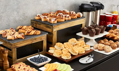 Continental breakfast catering spread including assorted baked goods such as cinnamon rolls, muffins, scones, croissants, and biscuits. Display also includes fresh fruit, coffee in pump pots, and juice. Wooden and slate catering risers. Assorted platters and red mugs.