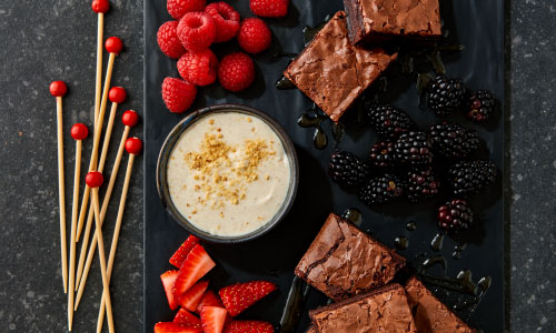Assorted berries and brownies assembled on a tray with creamy dip. Toothpicks with red wooden beads on the end in a pile on side of tray on dark granite counter.
