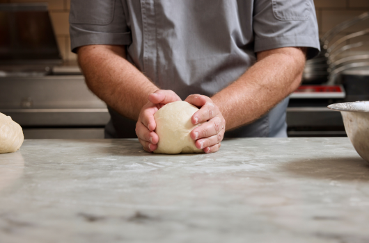 A person in a gray chef’s coat holding a dough ball on a countertop.