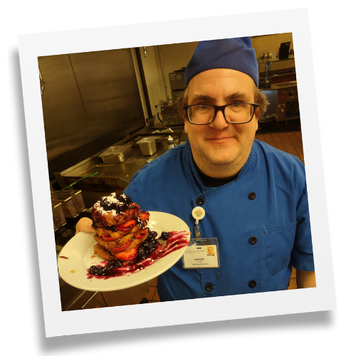 Man in blue chef jacket standing in a kitchen holding the Chocolate Berry Biscuit Bomb dessert creation on a white plate.