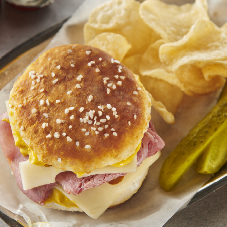 Top-down image of biscuit sandwich with ham, cheese, and mustard. Shown on parchment paper with chips and pickle spears.