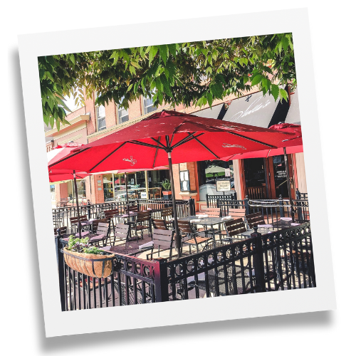 Red umbrella covering an outdoor, sunny patio, with a black fence bordering outdoor tables and chairs.