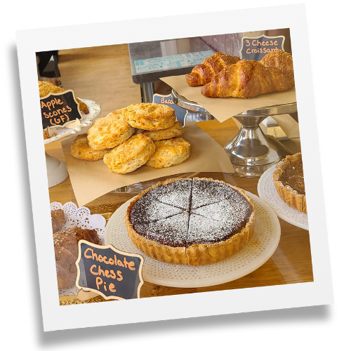 Chocolate chess pie and apple scone pastry signs displayed along with platters of biscuits, croissants, scones, and a chocolate cake covered in powder sugar.