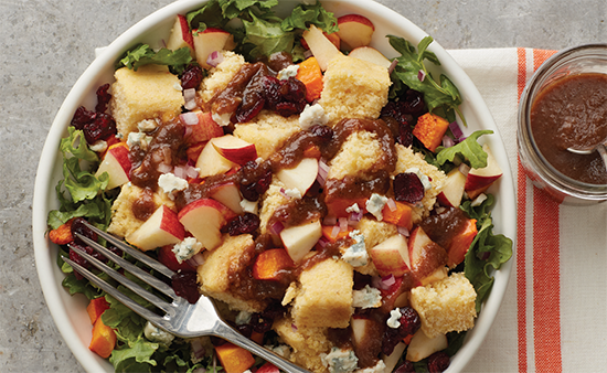Salad in a white bowl with greens, apples, cranberries and cornbread croutons sitting next to a napkin with dressing
